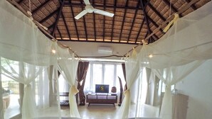 Une chambre avec deux lits recouverts de baldaquins blancs, un ventilateur de plafond et un téléviseur posé sur un meuble en bois.