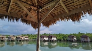 Des bungalows sur pilotis, avec toits de chaume, des piliers en bois et un plan d’eau calme dans lequel se reflètent les constructions.