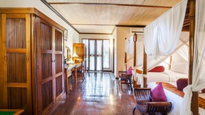 A room with a wooden floor, a bed with white bedding and a canopy, a wooden desk with a chair, and a large window with curtains. - Dwaraloka Retreat Ubud (Ubud)