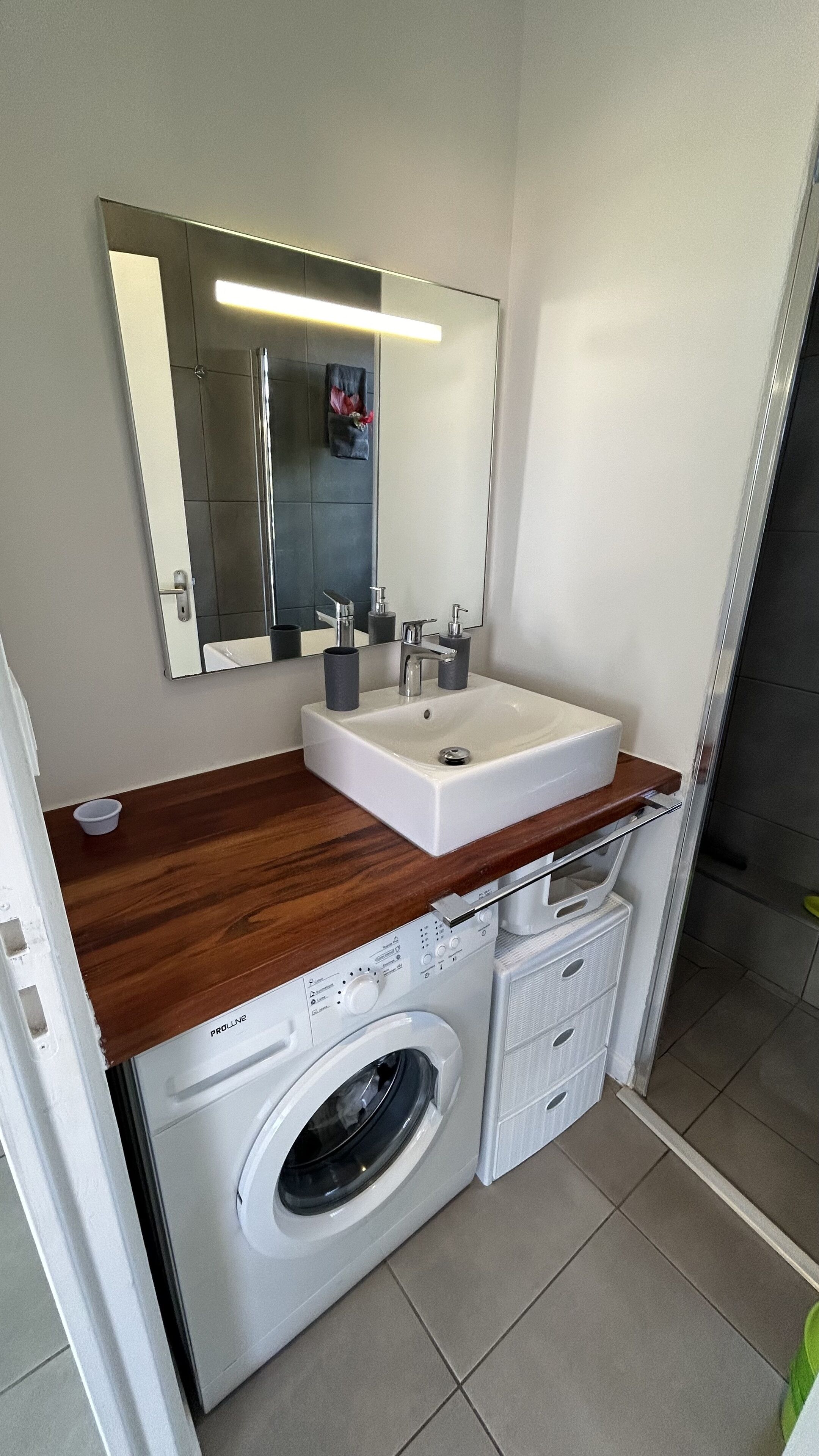 A compact bathroom with a white sink, a mirror, a wooden countertop, and a built-in washing machine.