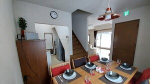 A dining area with a wooden table set for four, equipped with black bowls, plates, and cutlery. A clock and an emergency exit sign are visible on the walls. - Miyuki Townhouse (Furano)