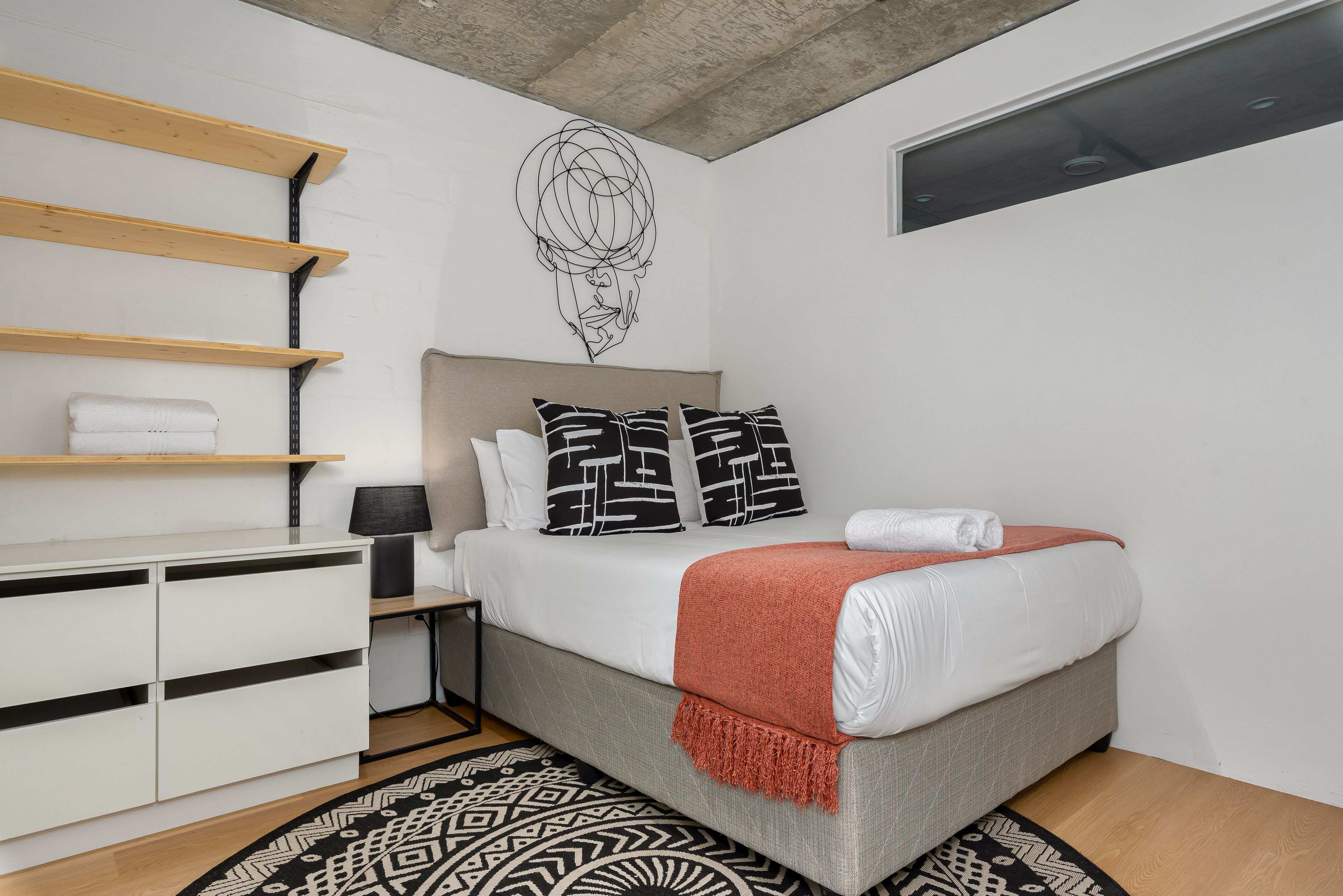 A modern bedroom with a bed, white dresser, shelves, and a patterned rug.