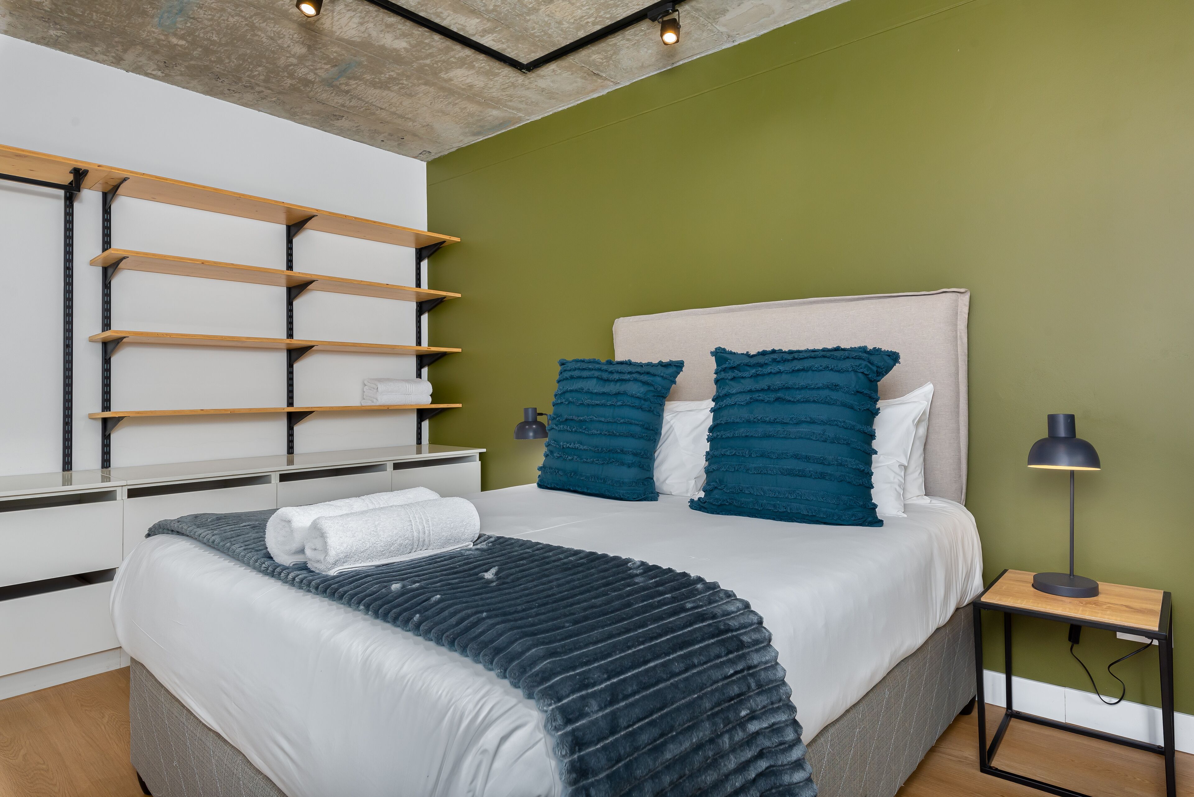 A neatly arranged bedroom with a bed, a bedside table with a lamp, and a wall-mounted shelf unit.