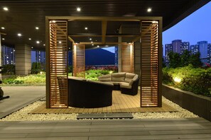 An outdoor lounge area with a wooden pergola, seating, and a view of the cityscape at dusk.