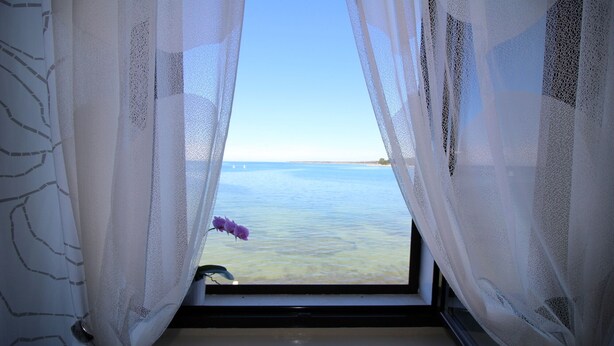 Une fenêtre ornée de rideaux légers, une vue sur la mer et une fleur violette posée sur le rebord de la fenêtre.