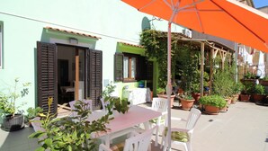 Une terrasse extérieure avec une table et des chaises, entourée de plantes en pot et agrémentée d’un parasol orange.