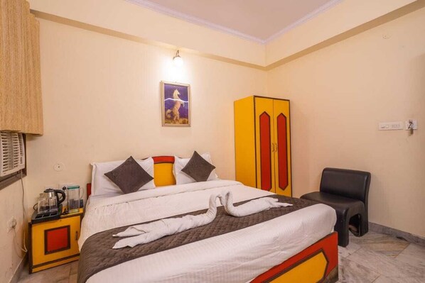 A hotel room with a bed, a wardrobe, a chair, and a small table with a kettle and a glass.