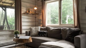 Een gezellig blokhutinterieur met een houten wand, een groot raam, een bank en een kleine tafel met een plant en documenten.