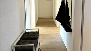 A narrow hallway with white walls, a wooden floor, and a ceiling light fixture. There is a coat rack with a black jacket hanging on it, and a small white shelf unit with a mattress on top. - Vacation apartment on a hillside with a picturesque view (Osterode am Harz)