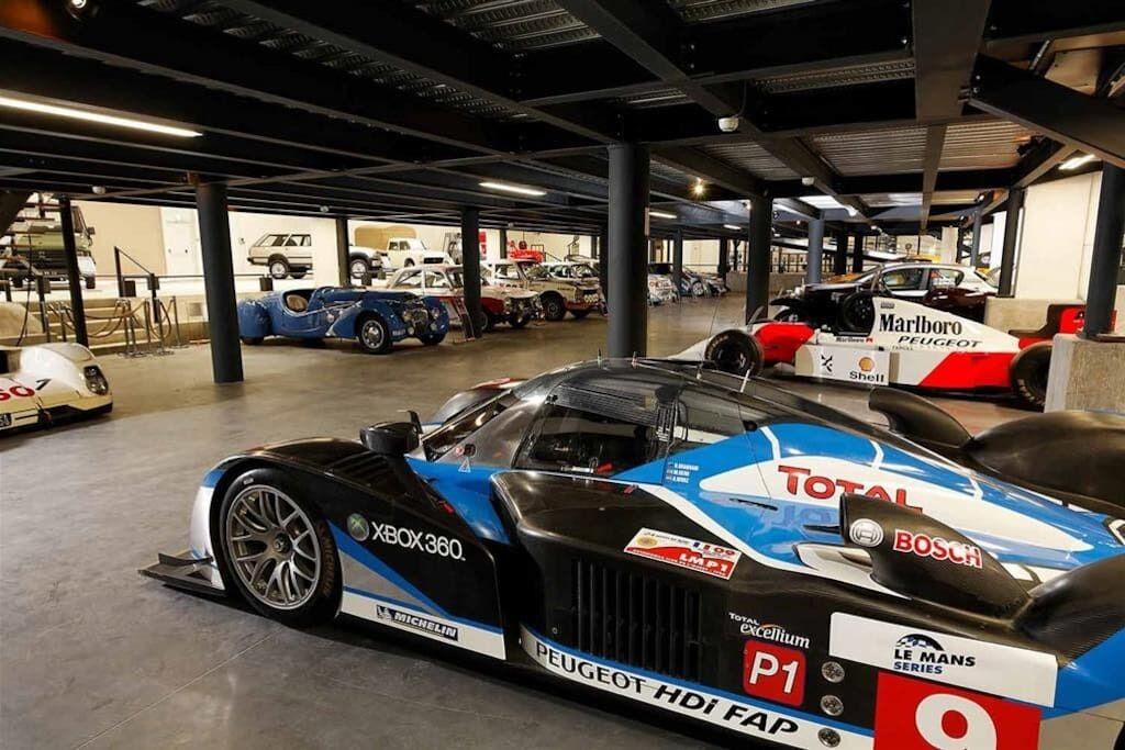 A racing car in a garage with other vehicles, indicating a motorsport setting.