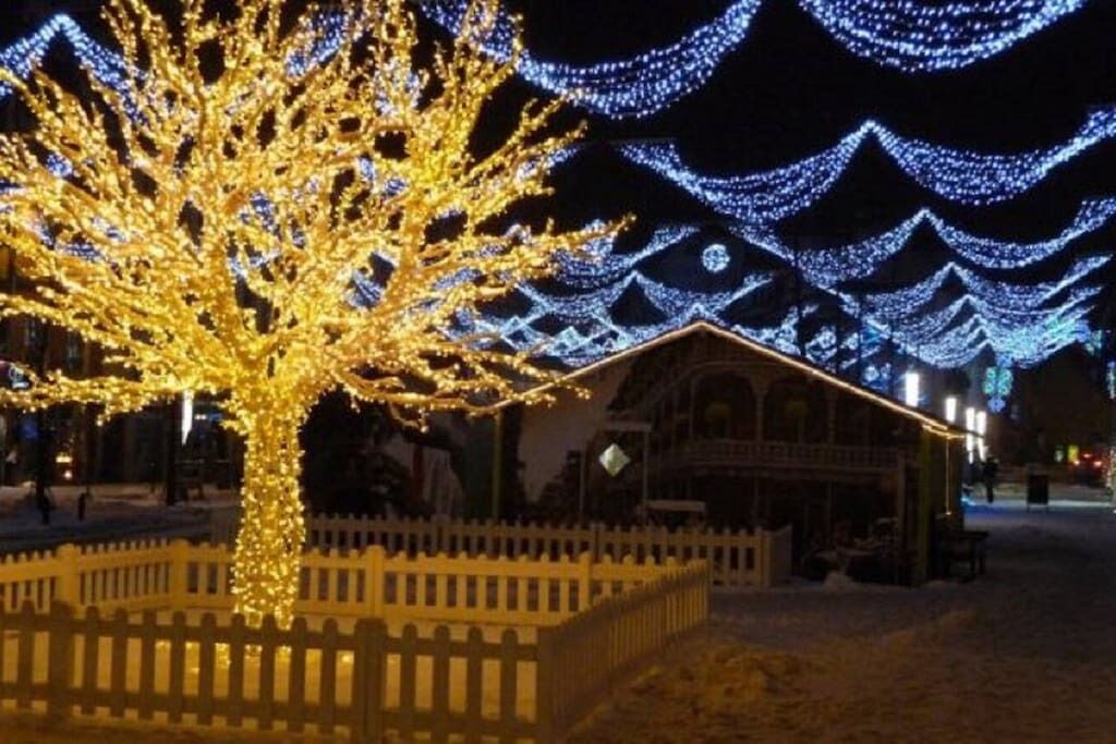 A snow-covered outdoor area with a tree decorated with lights, a wooden fence, and a building with a sloped roof.