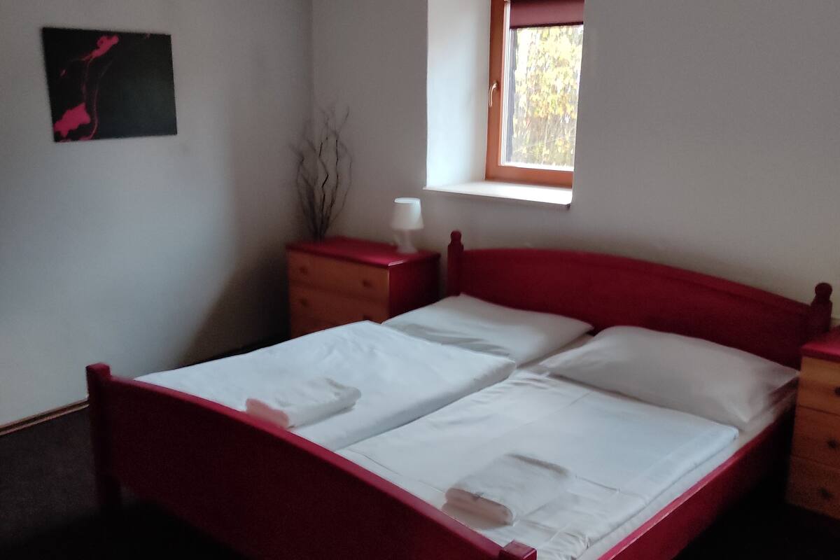 A bedroom with a red bed, a nightstand, a dresser, a window with a view of trees, and a painting on the wall.