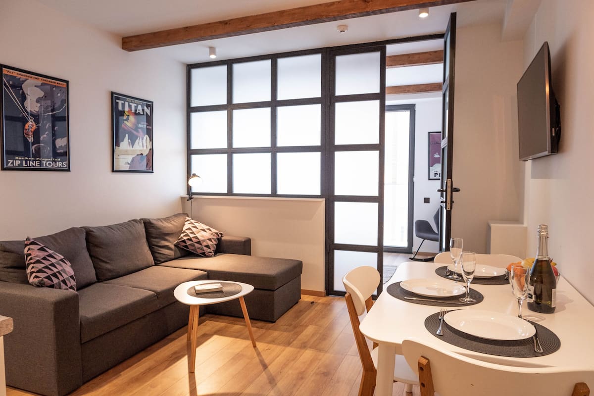A modern living room with a sofa, a dining table set for two, and a large window with blinds.