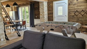 A cozy room with a stone wall, wooden ceiling, a sofa, a foosball table, a dining table with chairs, and a staircase. - Gîte Dune between orchard and Omaha beach (Colleville sur mer)