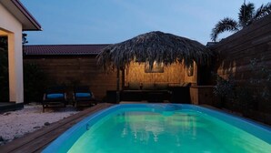 Una piscina amb una terrassa de fusta, una construcció amb teulada de palla i dues gandules.
