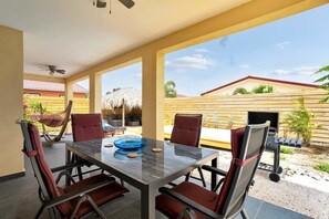 Un vaste espace extérieur aménagé pour les repas, avec une table en verre et des chaises rouges, donnant sur un jardin avec un barbecue et une structure à toit de chaume.