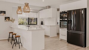 A modern kitchen with white cabinetry, a large island with stools, a black refrigerator, and pendant lighting. - The Little Distillery - Beachside Pool Retreat (Safety Beach)