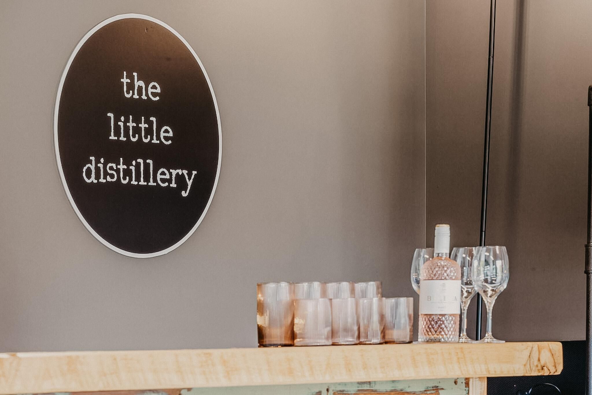 Una pared con un letrero que dice 'the little distillery' y una zona de bar con vasos y botellas.