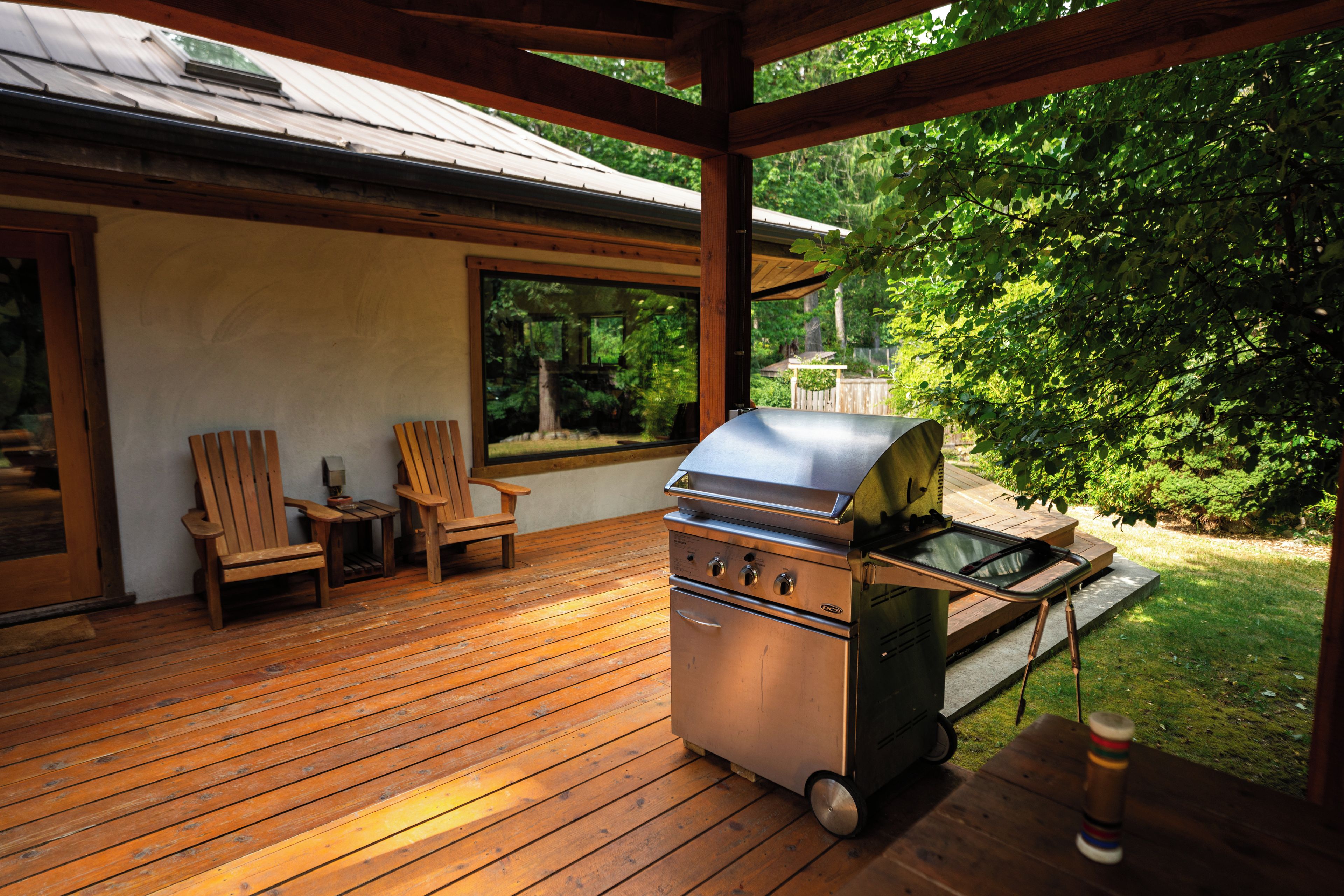 An outdoor patio with a grill, wooden deck, two chairs, and a view of a garden.