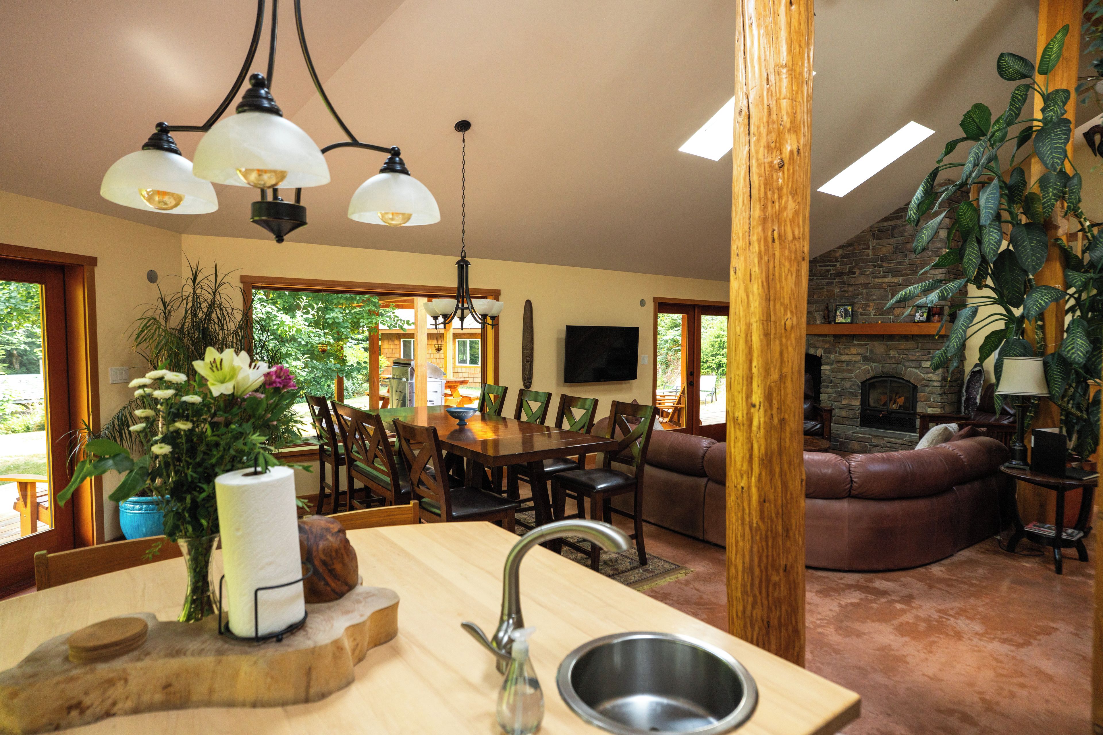 A well-lit dining area with a wooden table and chairs, a kitchen with a sink and a vase of flowers, and a stone fireplace in the living room.
