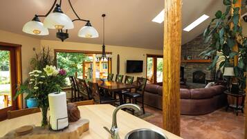 A well-lit dining area with a wooden table and chairs, a kitchen with a sink and a vase of flowers, and a stone fireplace in the living room.
