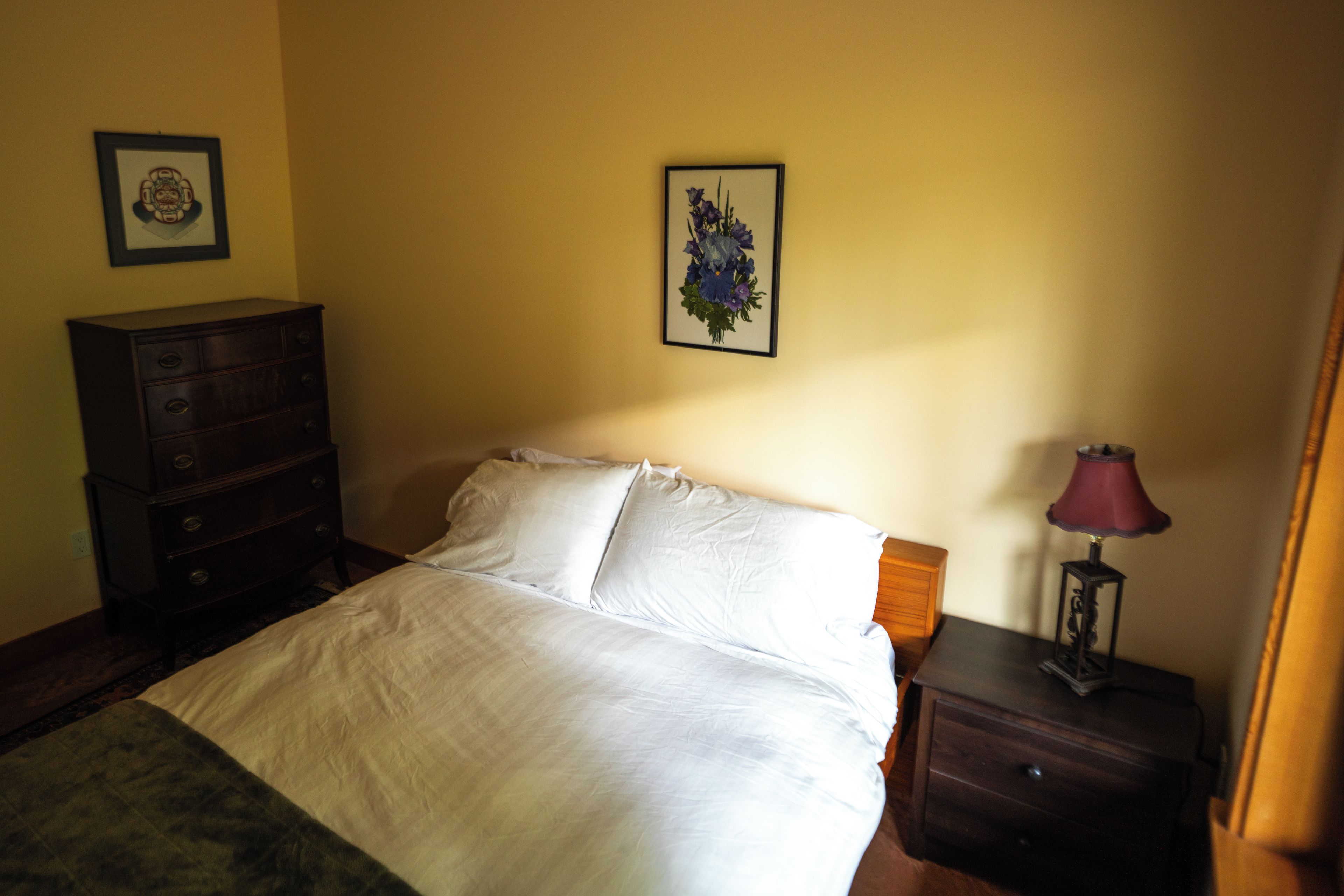 A bedroom with a bed, a dresser, a nightstand with a lamp, and two framed pictures on the wall.