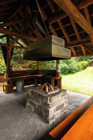 A rustic outdoor kitchen with a stone fire pit, wooden benches, and a hanging light fixture.