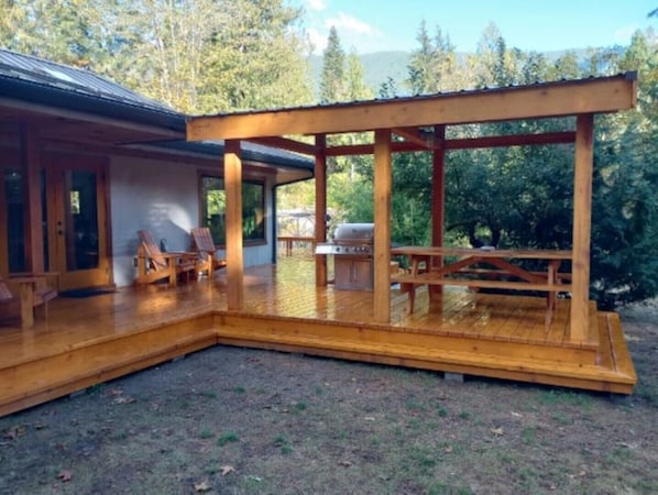 Una terraza de madera con zona de estar cubierta, una parrilla y vistas a un paisaje boscoso.