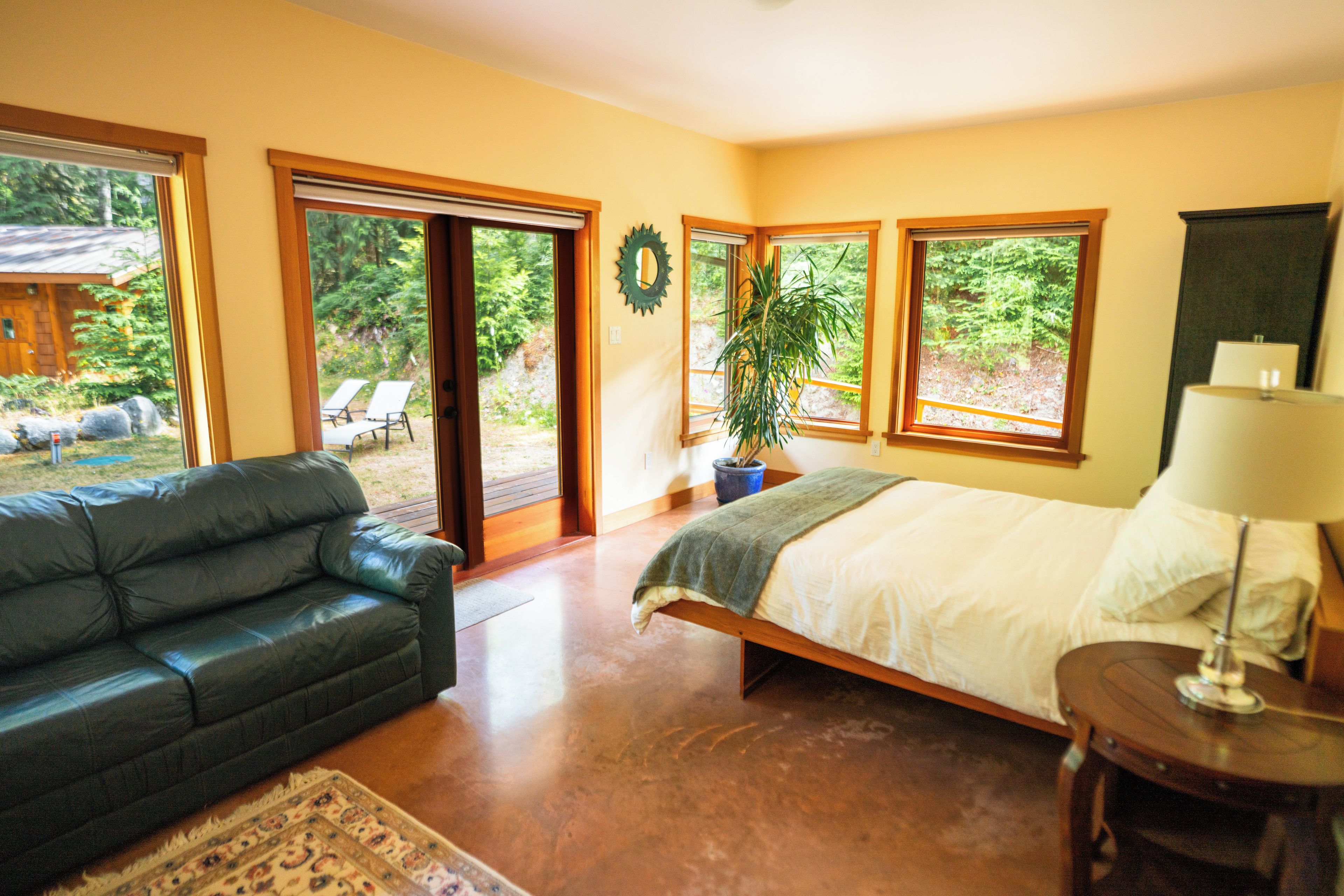 A bedroom with a bed, a nightstand, a lamp, and a view of the outdoors through large windows.