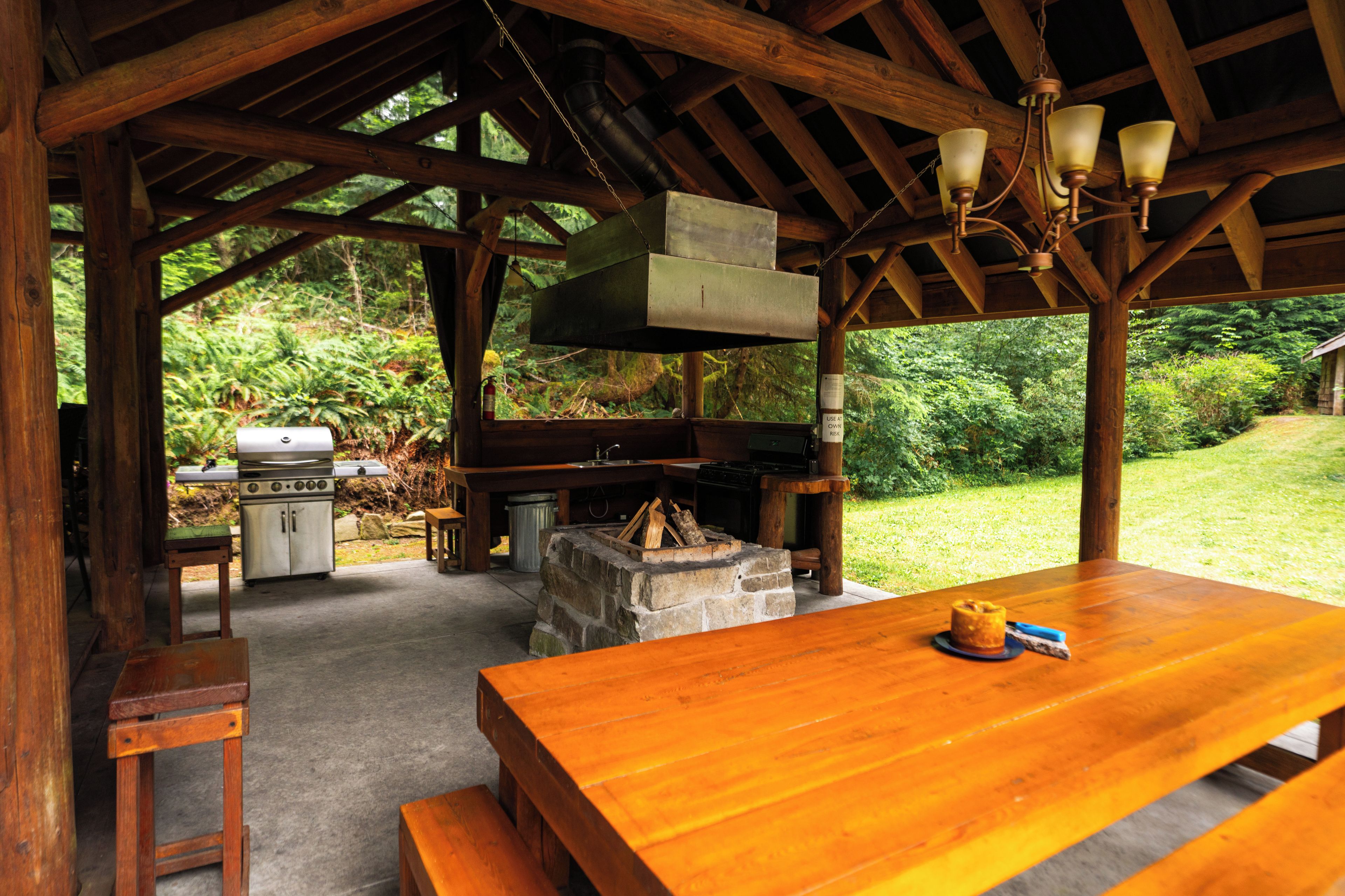 An outdoor wooden structure with a table, benches, a grill, and a fire pit.
