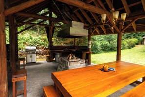 An outdoor wooden structure with a table, benches, a grill, and a fire pit.