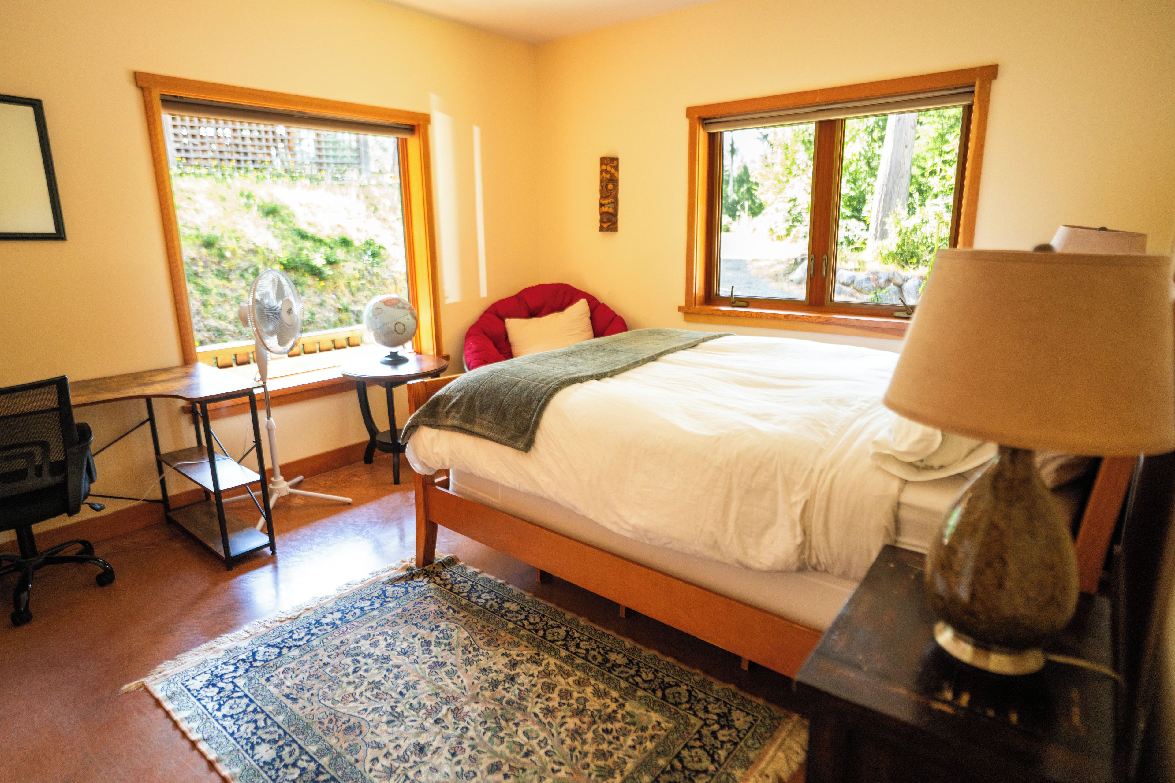 A bedroom with a bed, a desk, a chair, a lamp, and a window with a view of greenery.