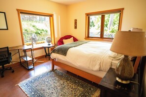 A bedroom with a bed, a desk, a chair, a lamp, and a window with a view of greenery.
