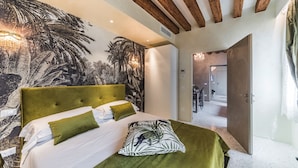 Une chambre avec un grand lit, une fresque représentant des palmiers, des poutres en bois au plafond et une vue sur une autre pièce.