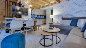 Un intérieur moderne comprenant un coin repas, une cuisine et un espace de vie, avec des poutres en bois, un grand téléviseur et un mur décoré sur le thème de la montagne.