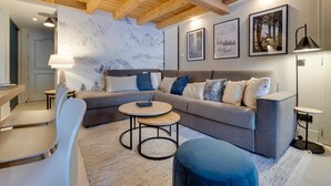 Un salon moderne avec un canapé gris, des coussins bleus et blancs, une table basse ronde et un pouf bleu. Le plafond est en bois avec des poutres apparentes, un mur est décoré de tableaux encadrés et une lampe sur pied est placée dans un coin.