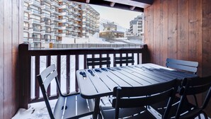 Un balcon en bois, agrémenté d’une table et de chaises, qui offre une vue sur une région montagneuse enneigée, avec un immeuble à plusieurs étages en arrière-plan.