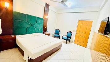 A bedroom with a bed, a ceiling fan, a green headboard, a wooden door, and a television on a wooden cabinet.