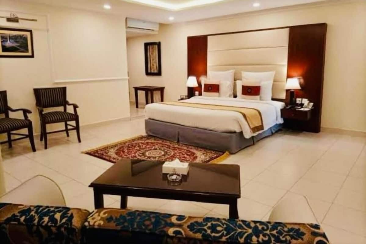 A hotel room with a bed, two chairs, a coffee table, a rug, and a nightstand with a lamp.