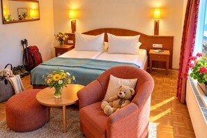 Habitación de hotel con una cama, dos mesitas de noche, una silla con un peluche, una pequeña mesa con un jarrón de flores y una ventana con cortinas.