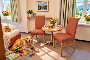 Una habitación acogedora con una cama, un perro, un oso de peluche, una mesa de comedor con sillas y una ventana con vistas.