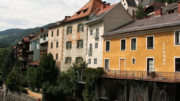 A row of traditional European buildings with balconies, a wooden fence, and a mountainous backdrop.
