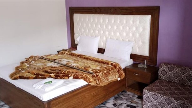 A neatly made bed with a patterned blanket, a wooden headboard, a nightstand with a lamp, and a patterned sofa. - Mezban Hotel & Restaurant (Balakot)
