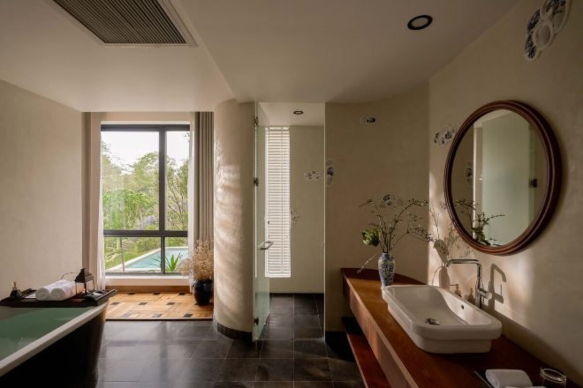 A modern bathroom with a large window, a wooden bench, a sink with a mirror, and a small plant.