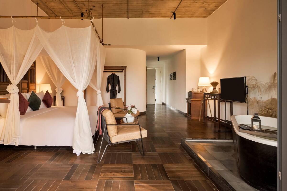 A bedroom with a canopy bed, a sitting area with a chair and table, a TV, and a bathroom with a bathtub.