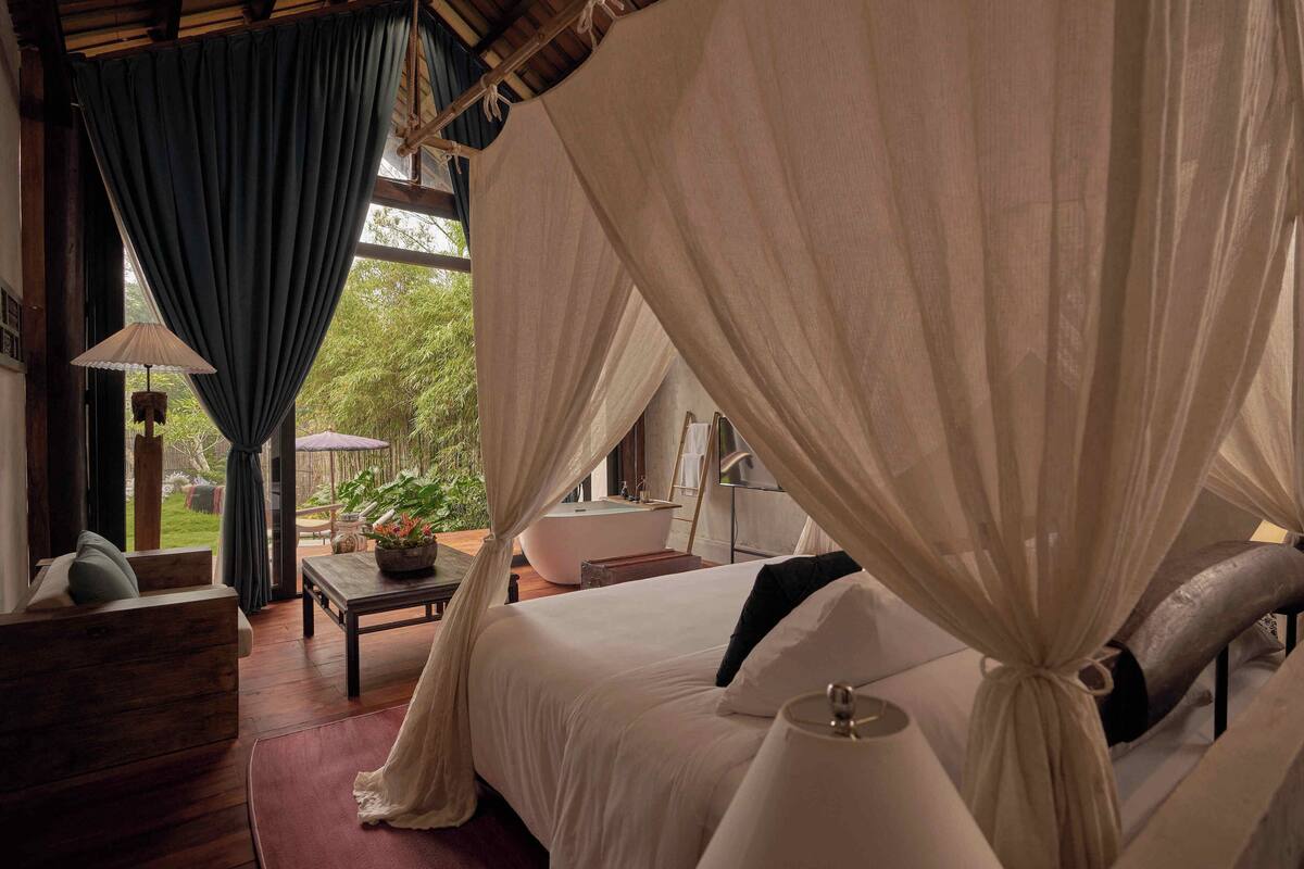 A bedroom with a canopy bed, a wooden nightstand, a table with a lamp, and a view of the outdoors.