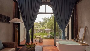 A bathroom with a freestanding bathtub, a wooden bench, a table with a vase of flowers, and a view of the garden through large windows. - aNhill Boutique (Hue)
