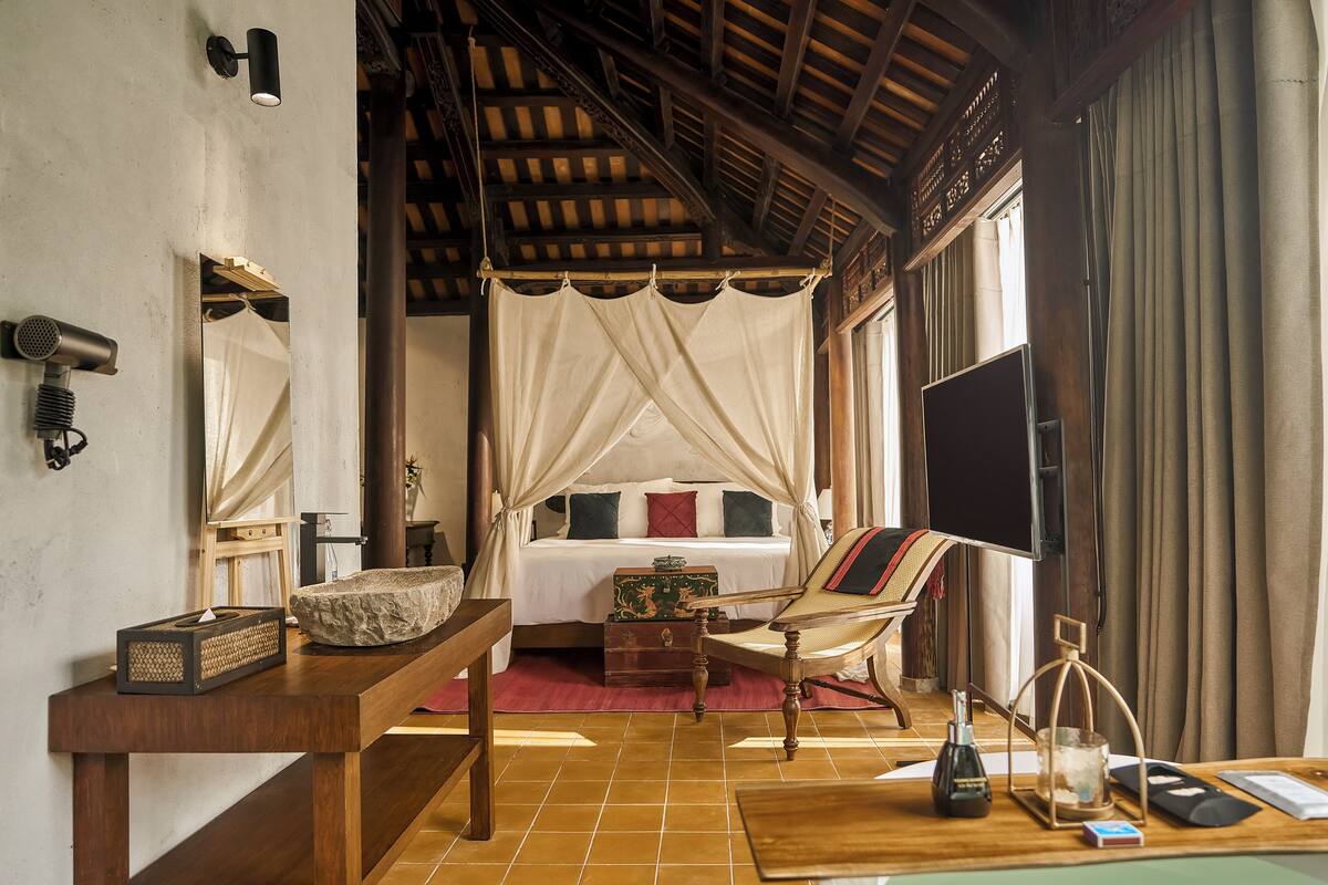 A bedroom with a canopy bed, a flat-screen TV, a wooden table with a vase, and a suitcase.