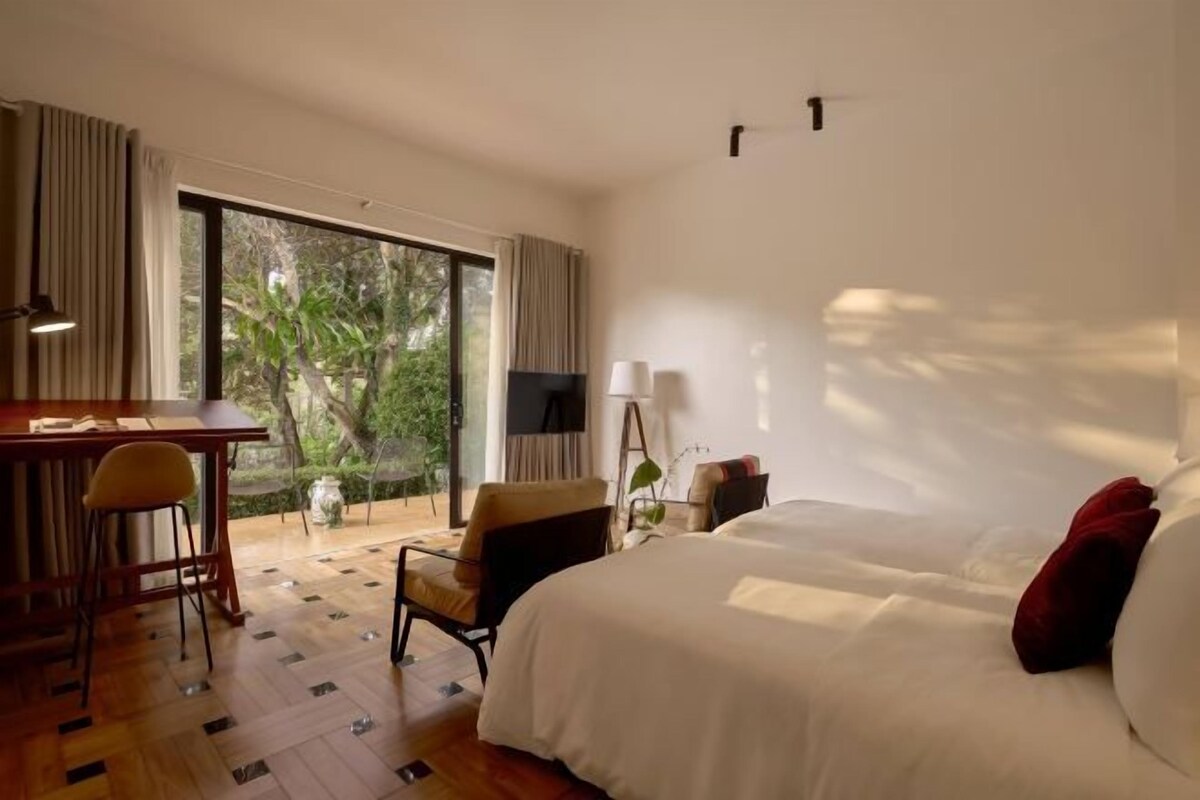A hotel room with a bed, a desk, and a chair. There is a large window with curtains, a TV, and a lamp.