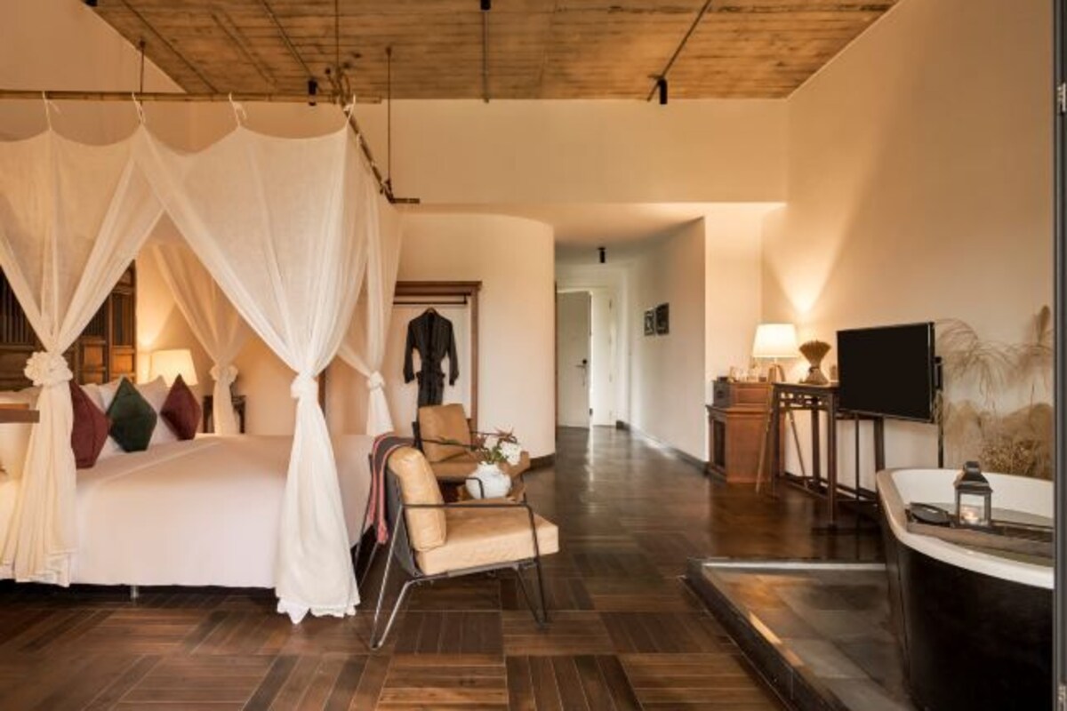 A bedroom with a canopy bed, a sitting area with a chair and table, and a TV on a stand.